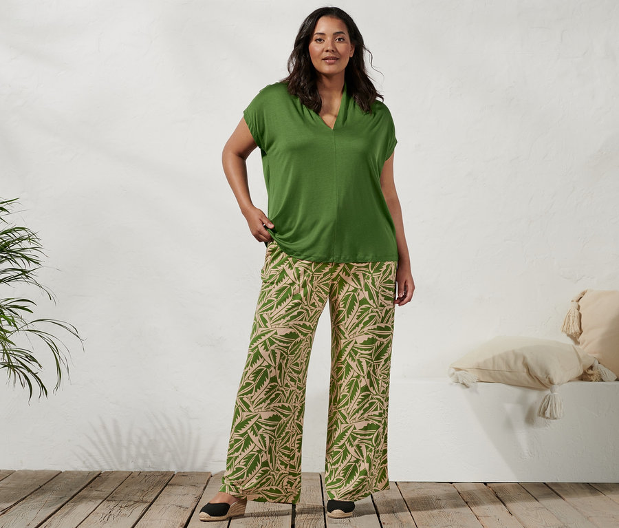 Žena stojí a má na sobě zelené triko se stojáčkem, zelenobéžové palácové kalhoty s potiskem a espadrilky s klínovým podpatkem.