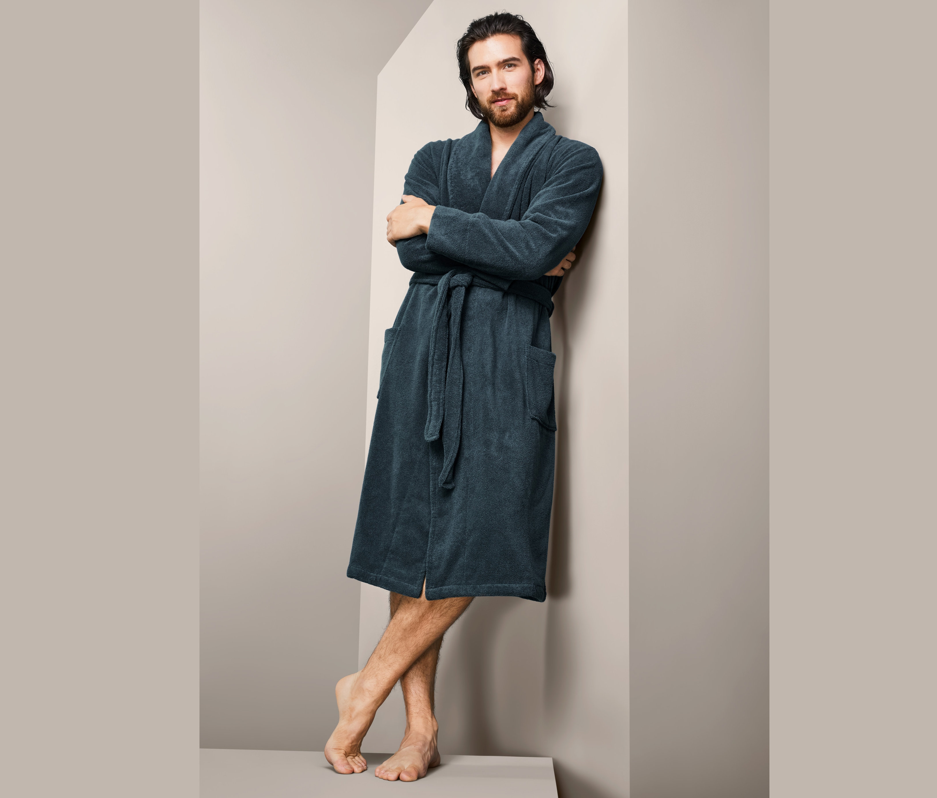 Muž s hnědými vlasy opřený o zeď v pánském županu.