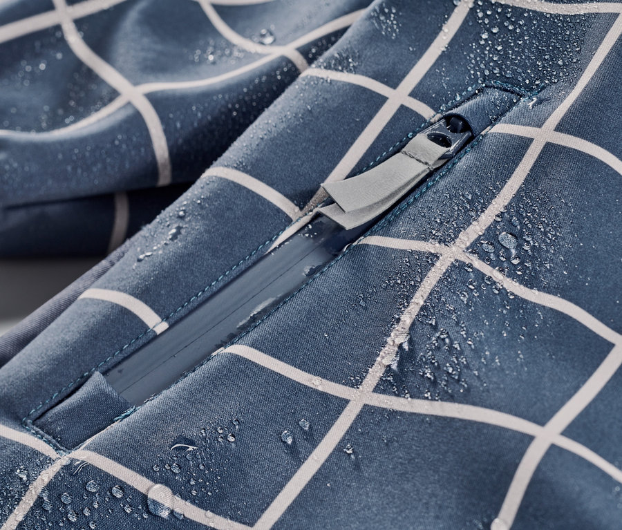 Detail dětské softshellové kombinézy s kostkovaným vzorem a zipem, na které jsou kapky vody.