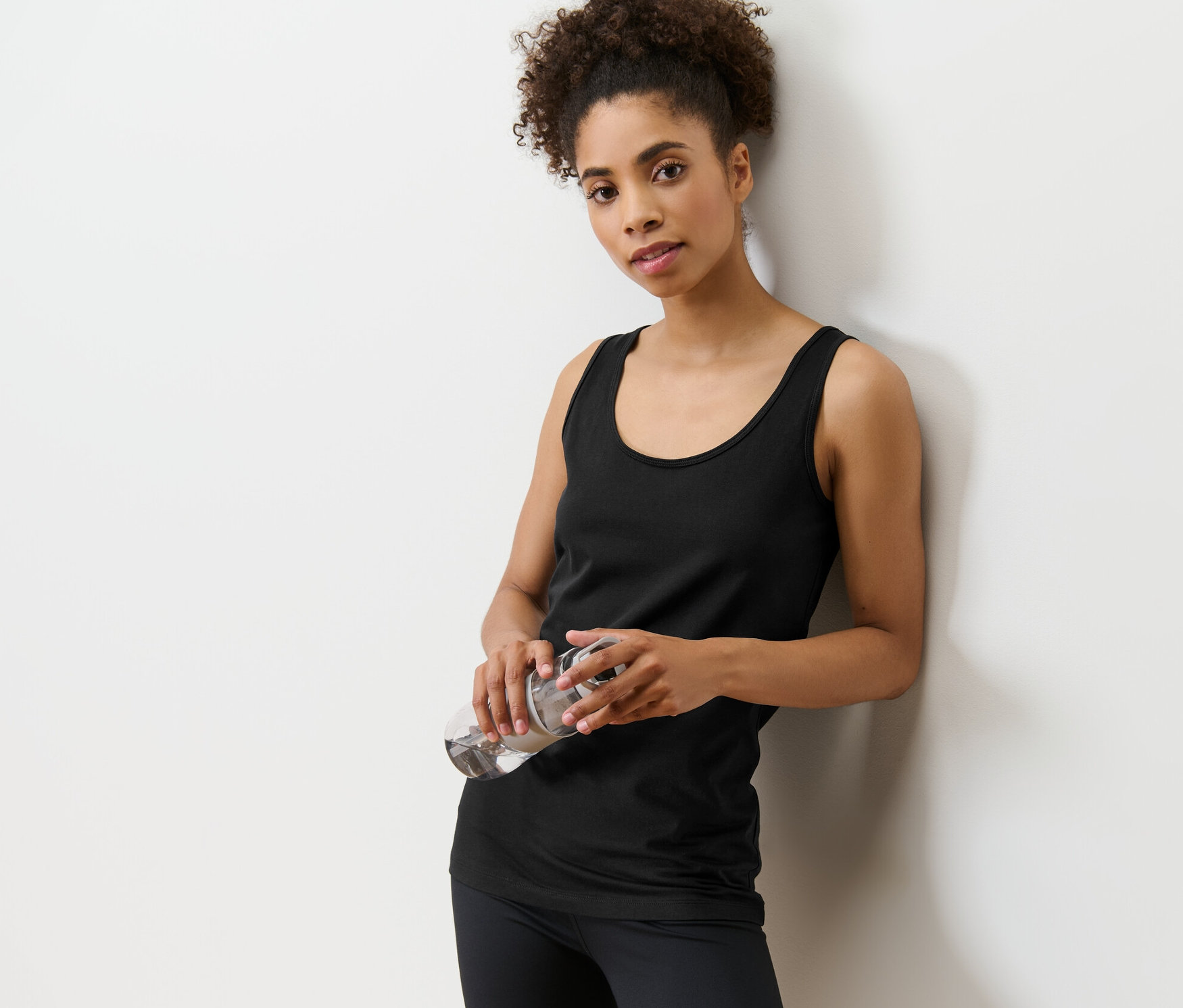 Žena opřená o zeď drží v rukou láhev s vodou. Má na sobě černé tílko z dvoudílné sady topů.