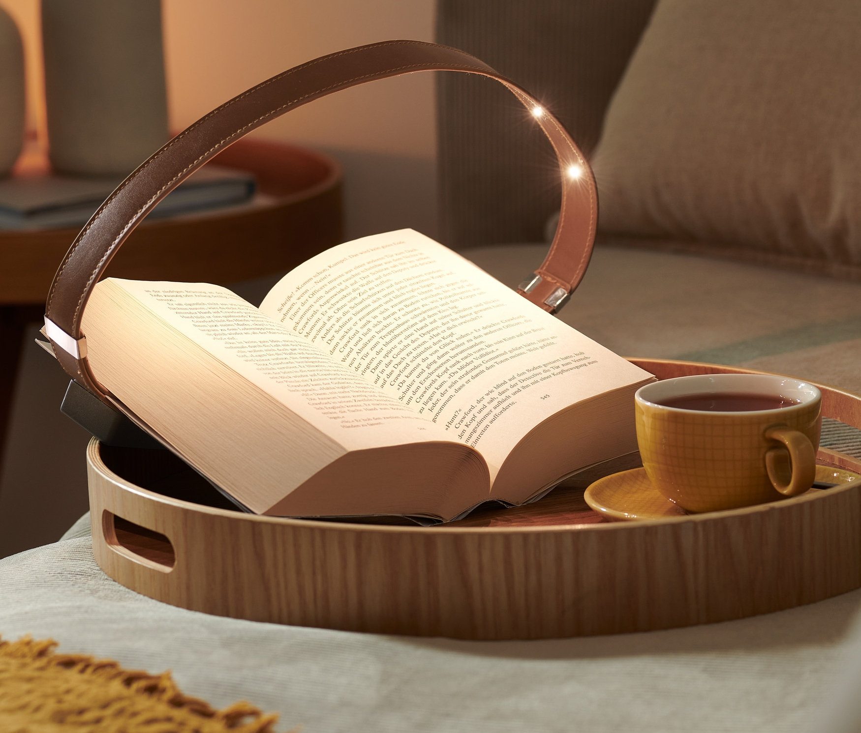 Otevřená kniha leží na dřevěném podnosu s lampičkou na čtení s magnetickým klipem a šálkem čaje.