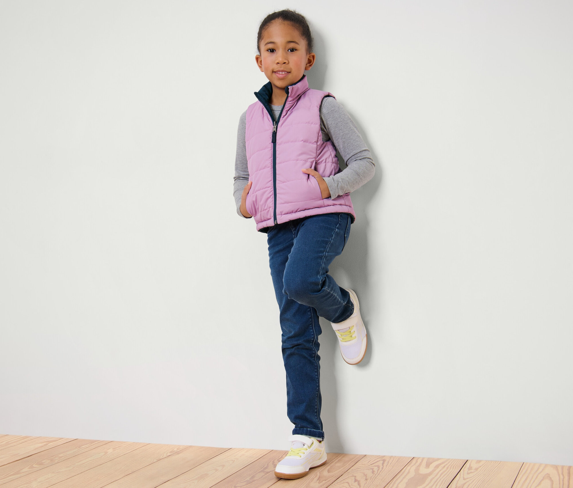 Dívka opřená o stěnu má na sobě dětskou oboustrannou prošívanou vestu, džíny a tenisky.