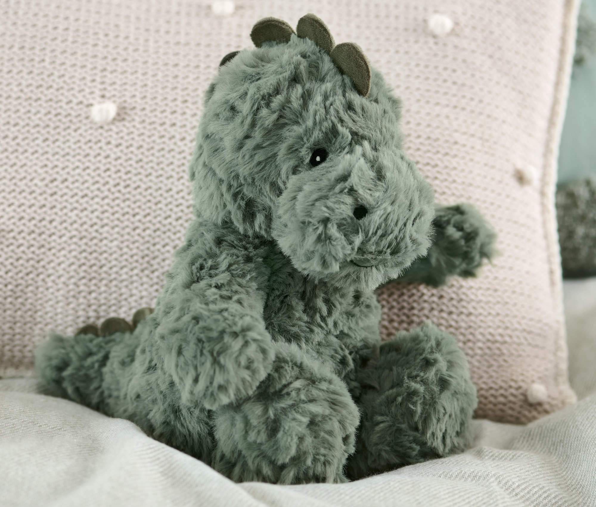 Plyšový dinosaurus sedí na posteli vedle béžového polštáře.