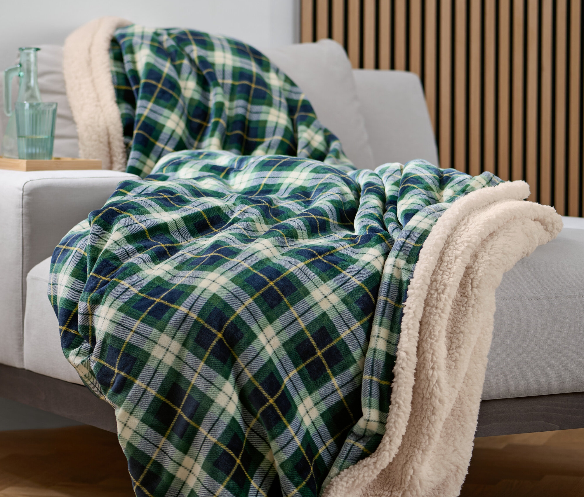 Zelená kostkovaná plyšová deka leží na šedé pohovce. Na bočním stolku stojí skleněná karafa a sklenice.