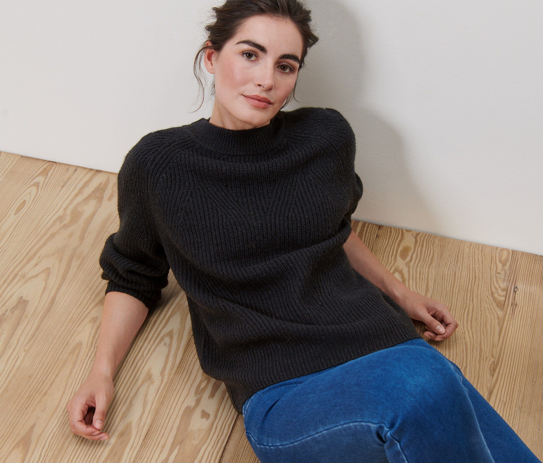 Žena leží na dřevěné podlaze a má na sobě černý pletený svetr.