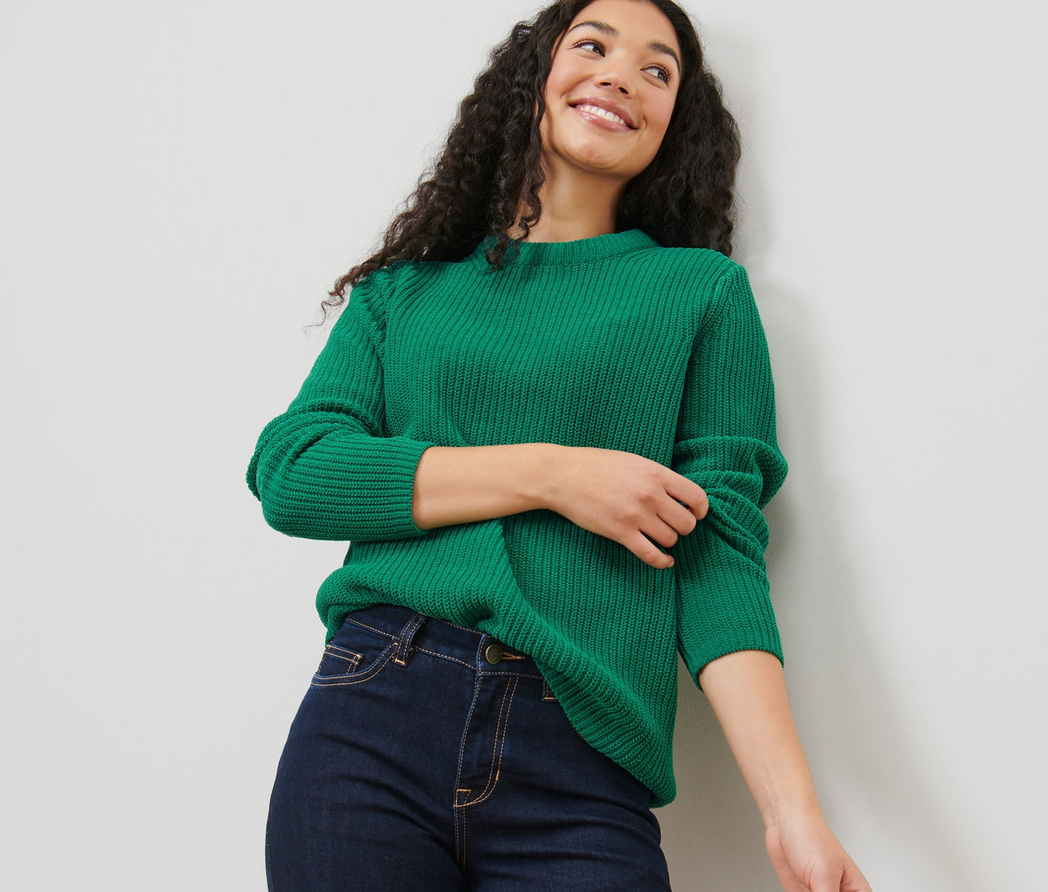 Žena s tmavými kudrnatými vlasy má na sobě zelený svetr z hrubého úpletu a džíny.