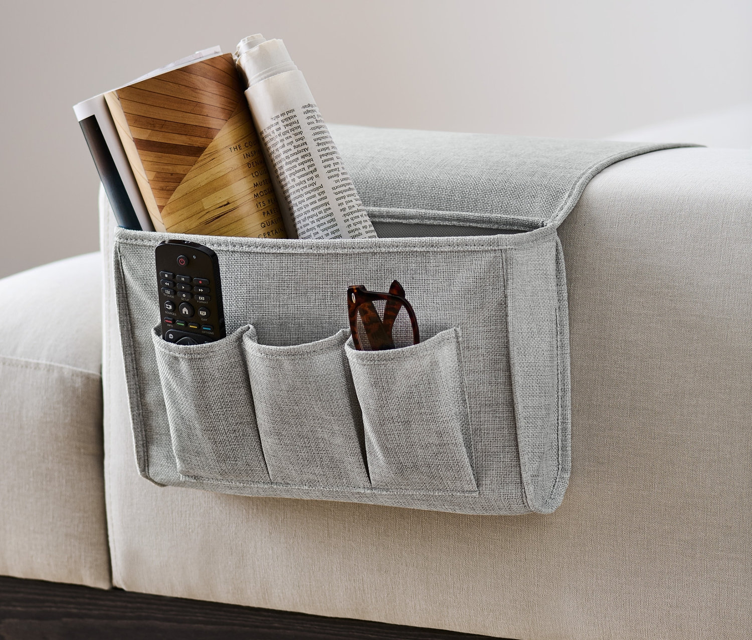 Šedý organizér na postel a pohovku visí na boku pohovky a obsahuje dálkový ovladač, brýle, knihy a noviny.