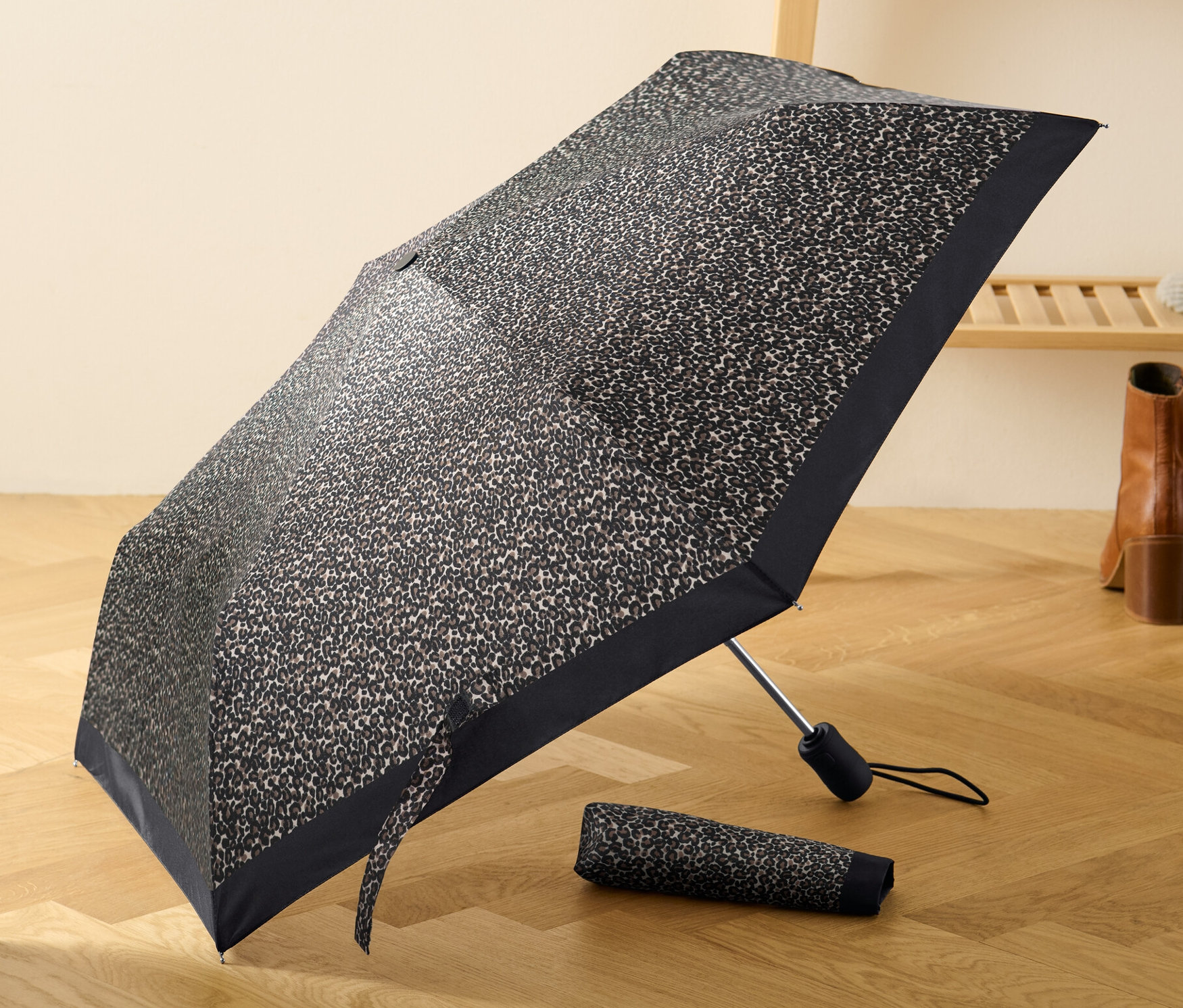 Otevřený automatický skládací deštník s leopardím vzorem leží na dřevěné podlaze vedle složeného deštníku.