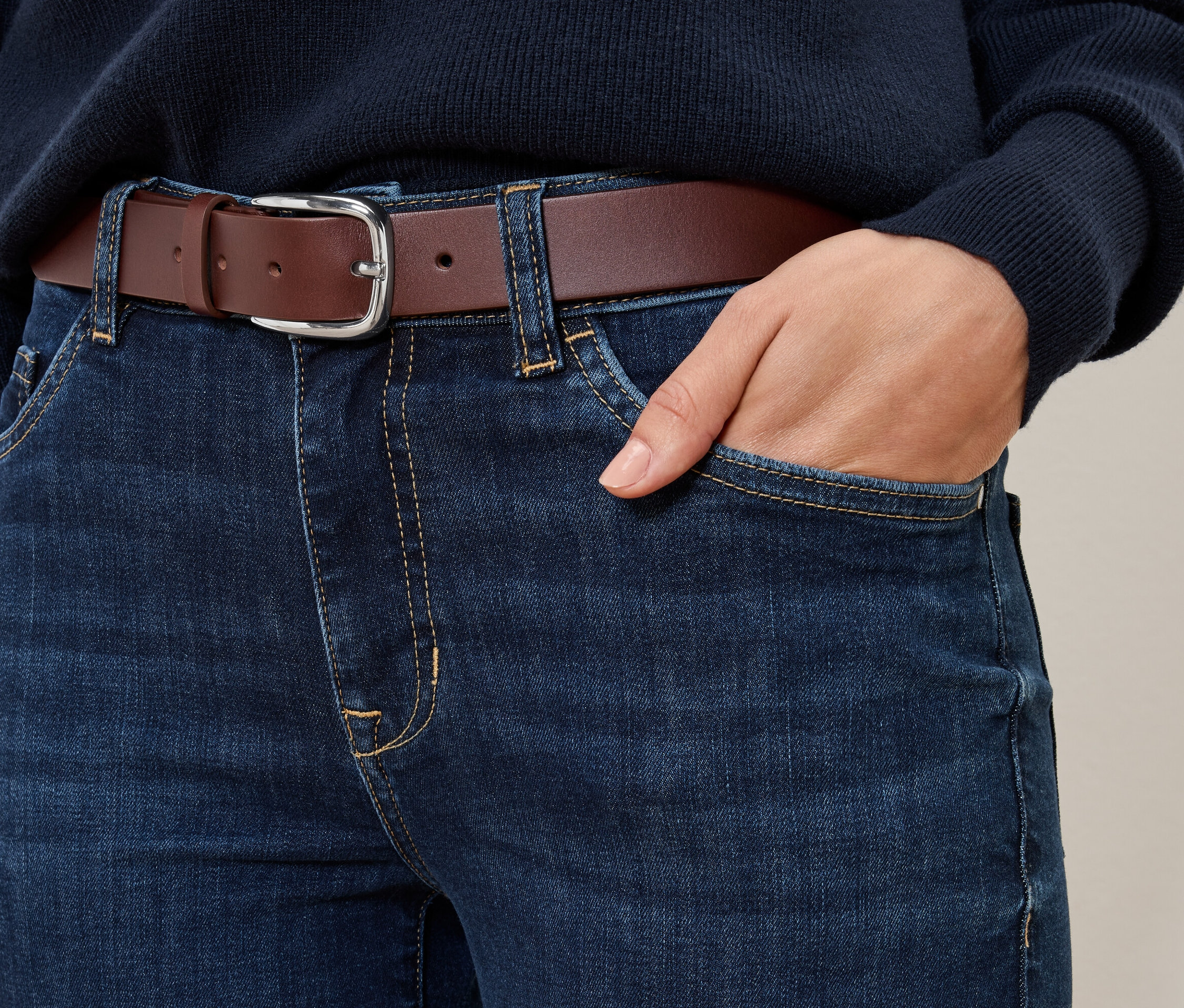 Detail džínsů s koňakovým koženým opaskem a rukou v kapse.