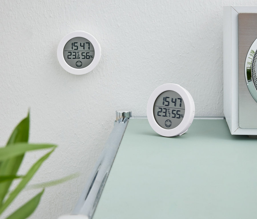 Dva bílé digitální hodiny. Jedny hodiny jsou na zdi. Druhé hodiny stojí na stole vedle bílého rádia.