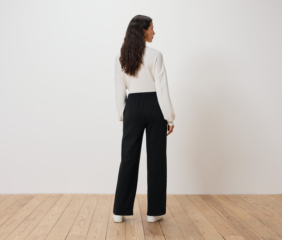 Žena v bílém svetru a černých kalhotách stojí zády k divákovi.