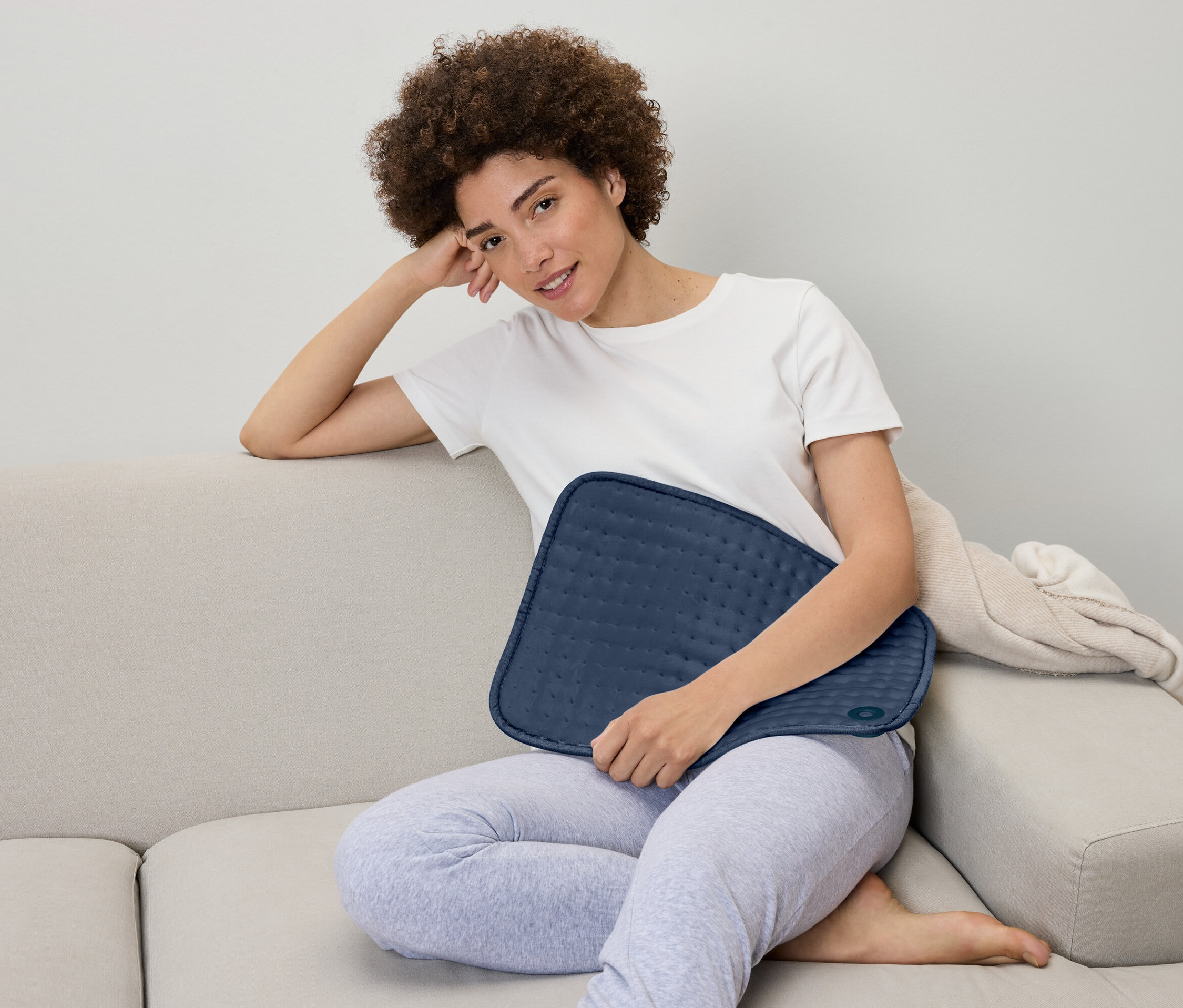 Žena sedí na gauči a drží na klíně modrý vyhřívací polštář.