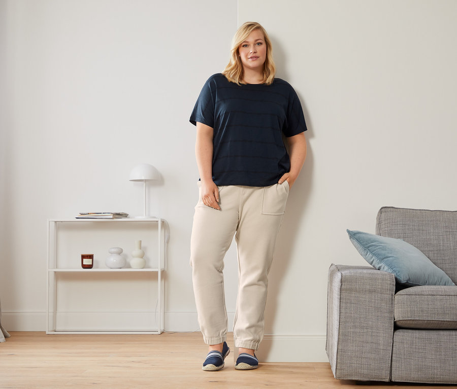 Žena opřená o zeď má na sobě tmavě modré triko s kulatým výstřihem, joggingové kalhoty v délce po kotníky a espadrilky.