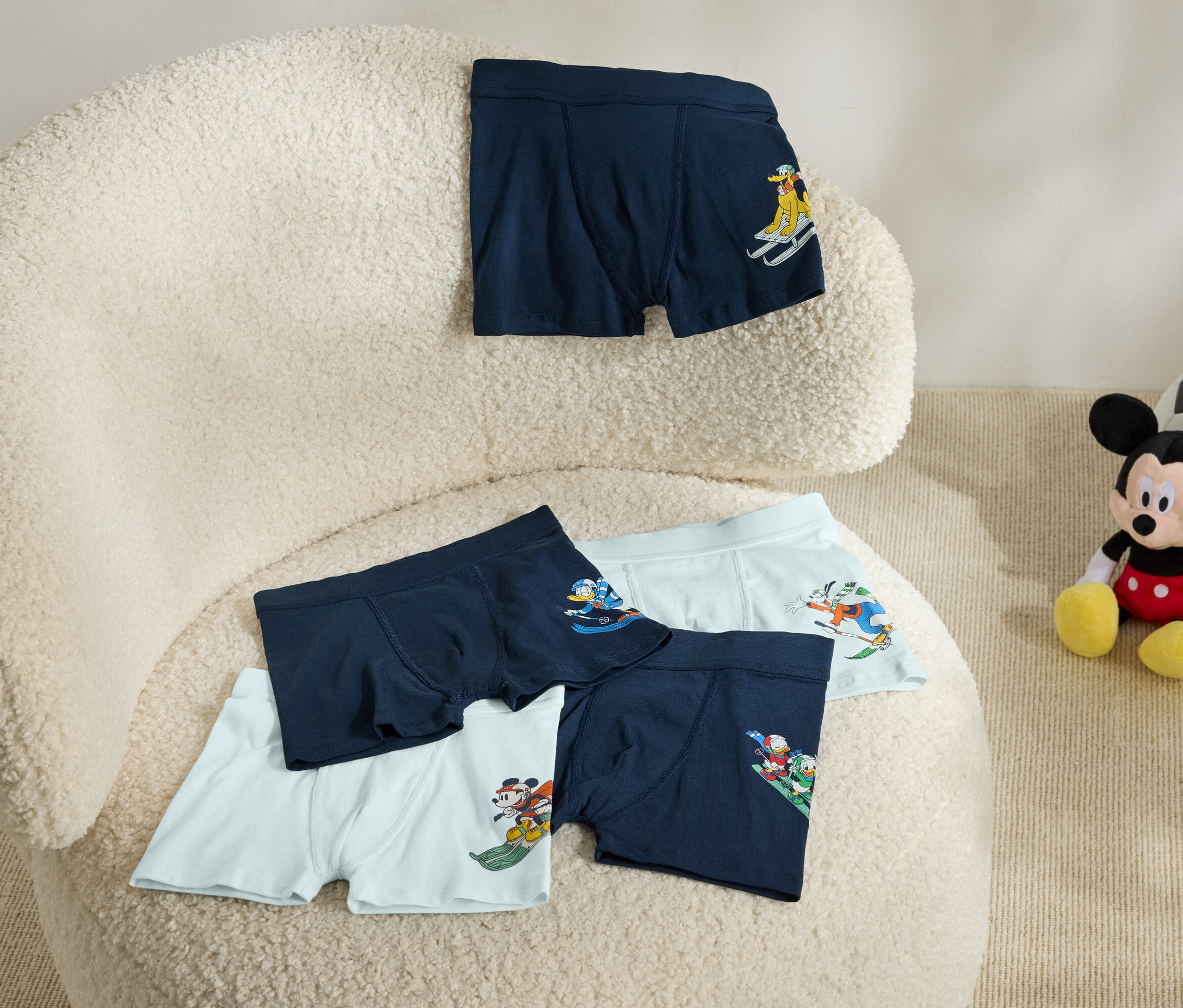 Na béžovém křesle leží pětery boxerky s potiskem. Vedle křesla sedí plyšová hračka Mickey Mouse s fotbalovým míčem.