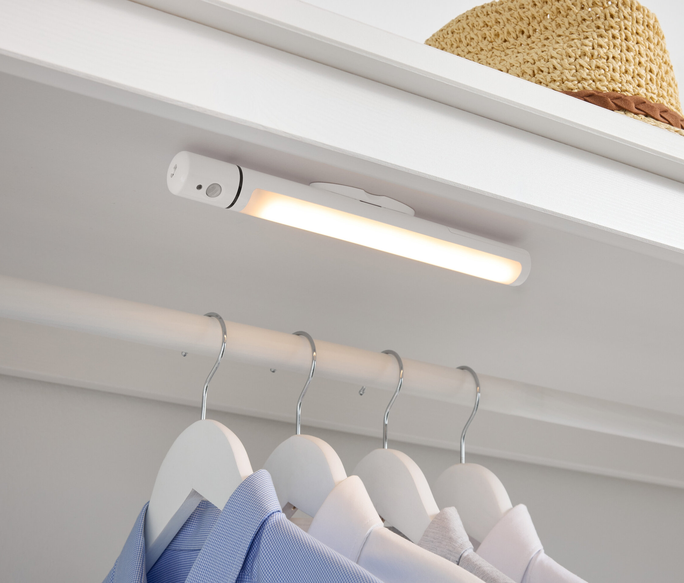 V šatníku je bílá LED lampa, pod ní visí na bílých ramínkách košile a nahoře leží slaměný klobouk.