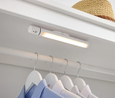 V šatníku je bílá LED lampa, pod ní visí na bílých ramínkách košile a nahoře leží slaměný klobouk.