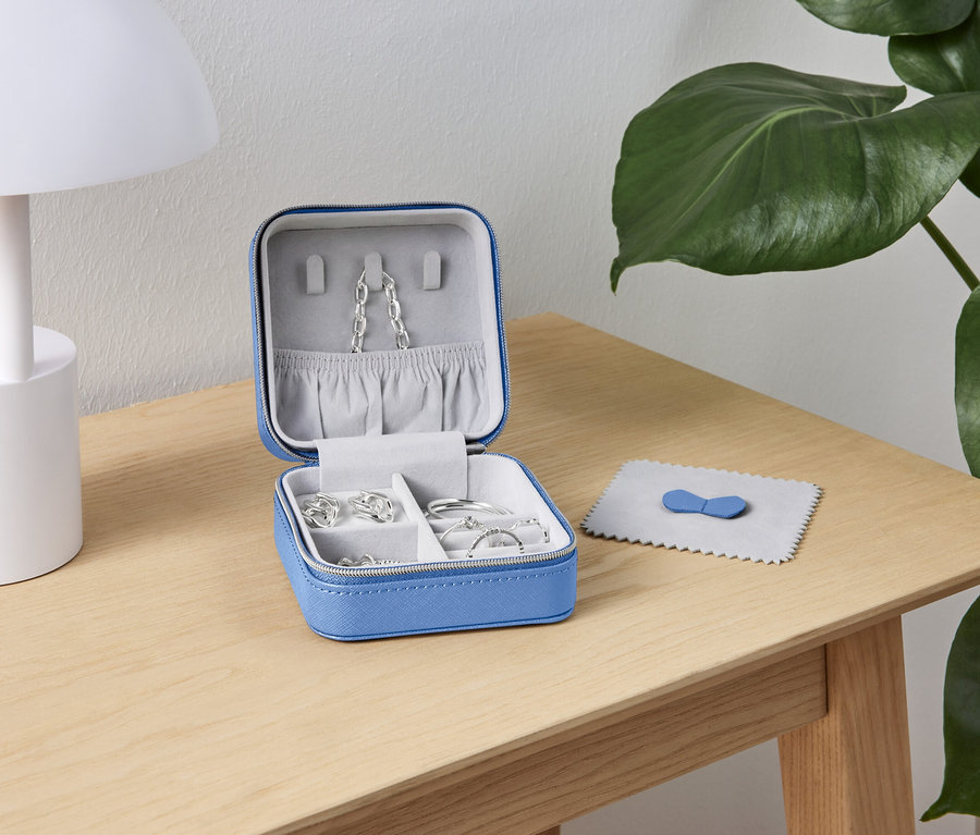 Otevřená modrá šperkovnice se stříbrnými šperky stojí na dřevěném stole vedle bílé lampy a zelené rostliny.