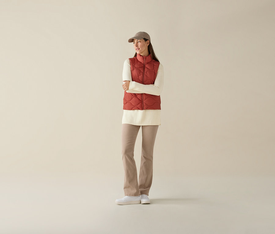 Žena se zkříženýma rukama na sobě má prošívanou vestu, krémový top, béžové kalhoty a kšiltovku.