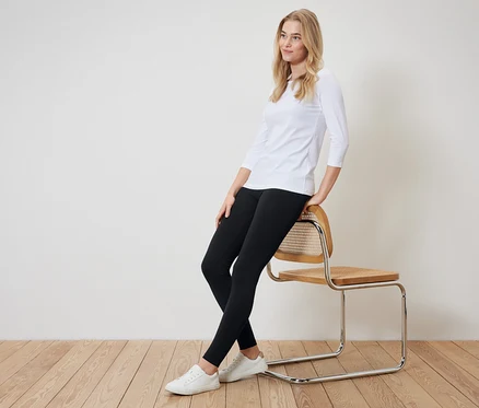 Blondýna sedí bokem na židli. Má na sobě bílé tričko s tříčtvrtečním rukávem, černé legíny a bílé tenisky.