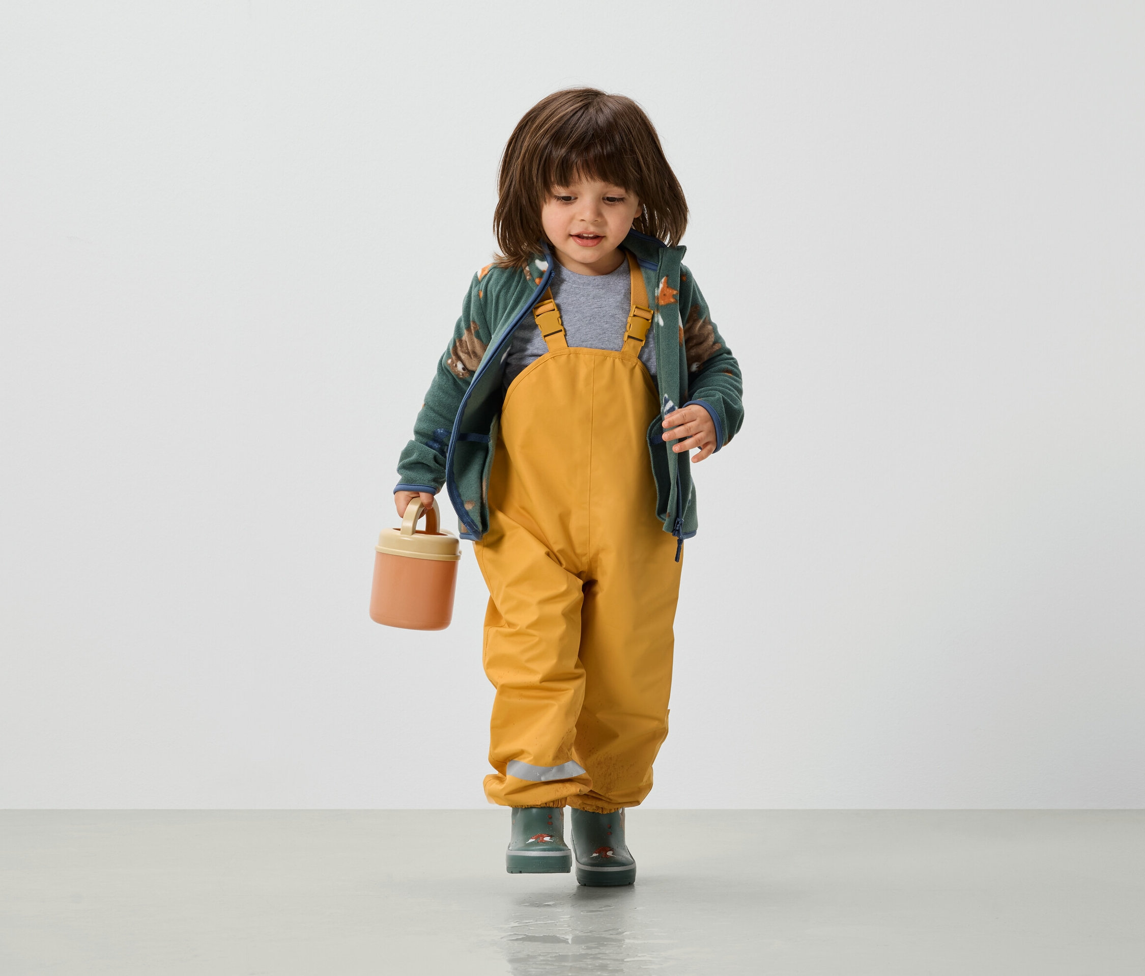 Batole v žlutých kalhotách s laclem, zelené bundě a holínkách drží v ruce oranžový box na svačinu.