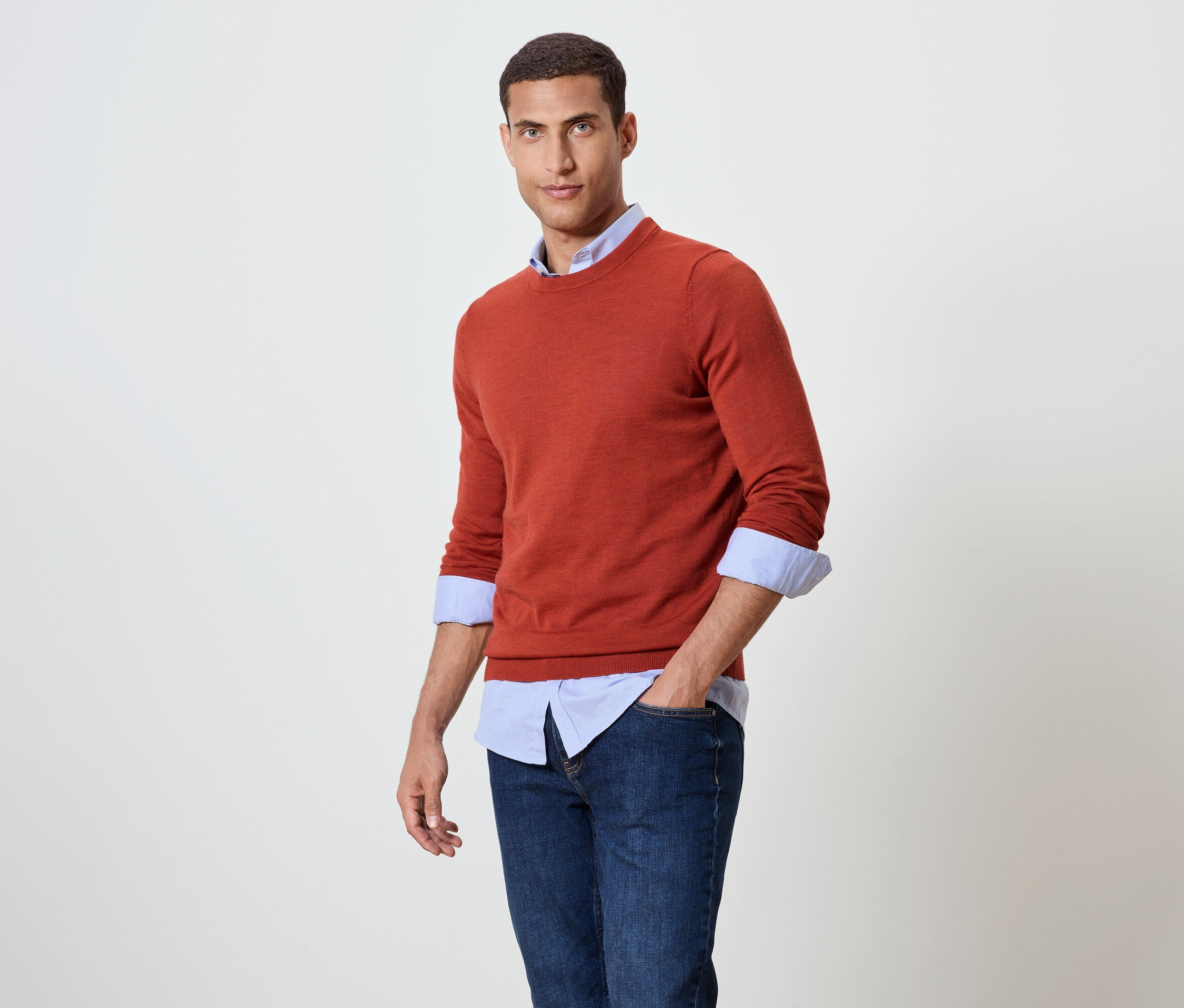 Muž pózuje v oranžovém svetru přes modrou košili a džíny.