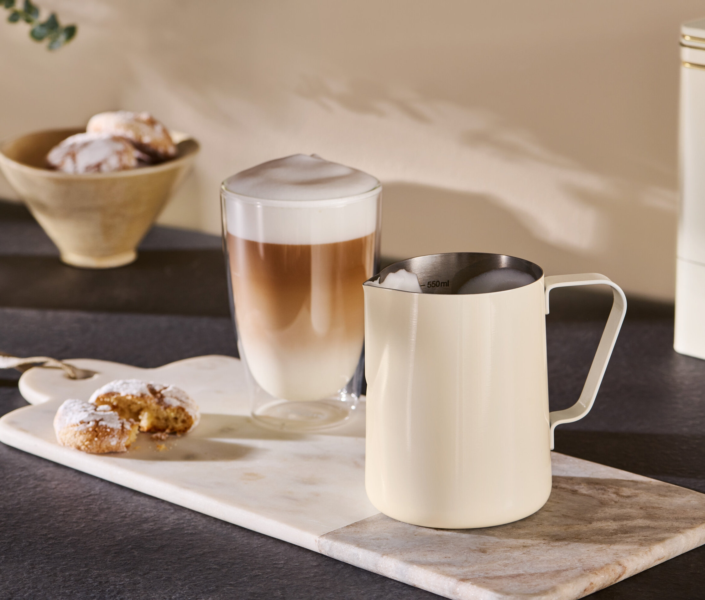 Béžová konvička na mléko stojí vedle sklenice s kávou na mramorovém podnosu.