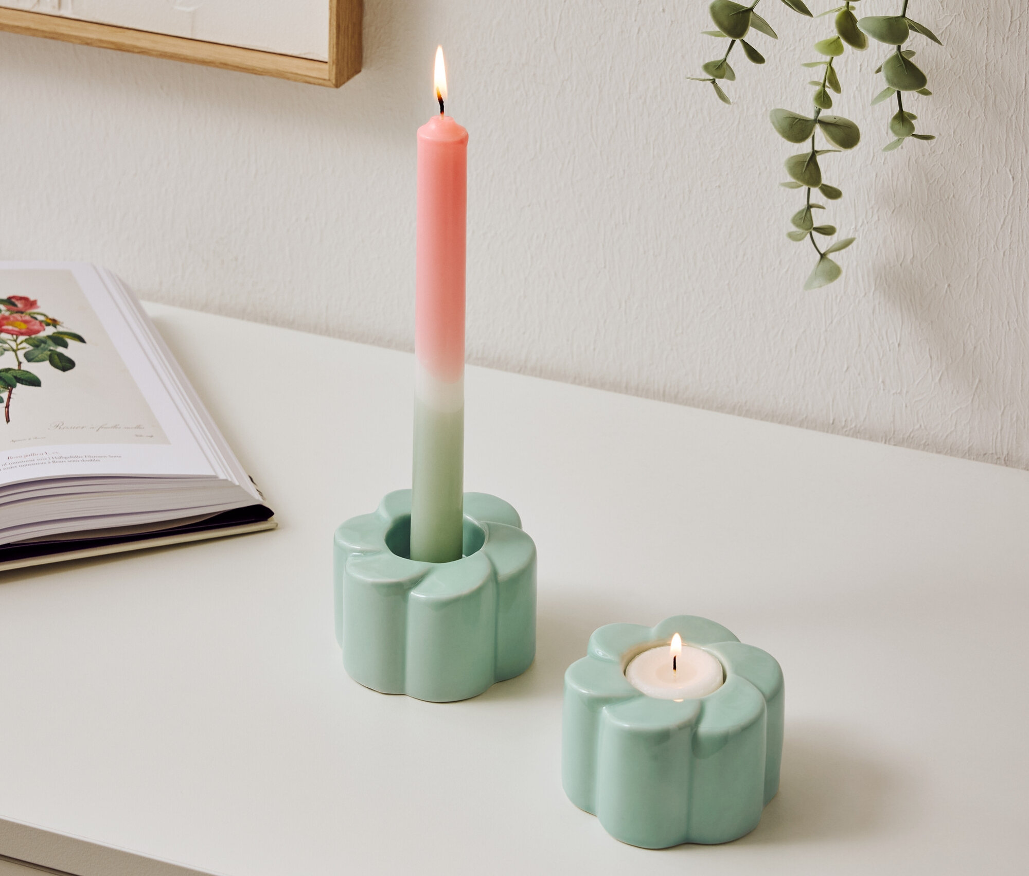 Dva světle modré svícny ve tvaru květin stojí na stole, jeden s hořící dvoubarevnou svíčkou.