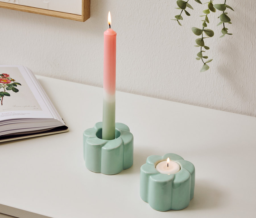 Dva světle modré svícny ve tvaru květin stojí na stole, jeden s hořící dvoubarevnou svíčkou.