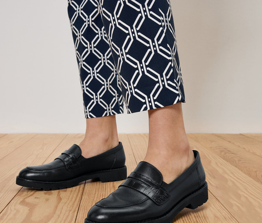 Nohy osoby v černých mokasínách a modrých vzorovaných kalhotách stojící na dřevěné podlaze.