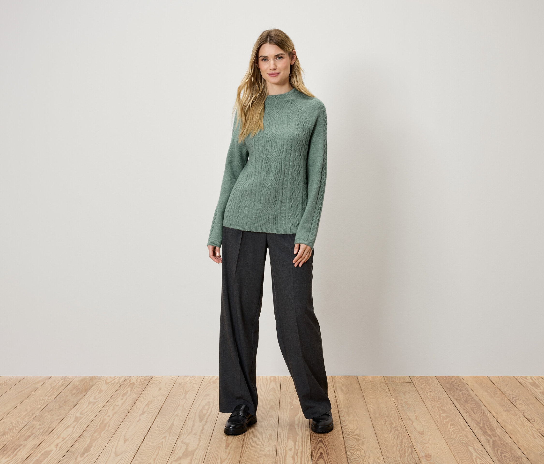 Blondýna stojí na dřevěné podlaze a má na sobě zelený pletený svetr a černé kalhoty.