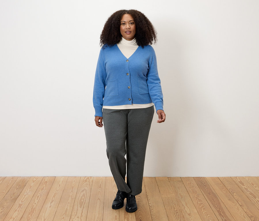 Žena v krémově bílém triku s rolákem, modrém svetru a šedých kalhotách.