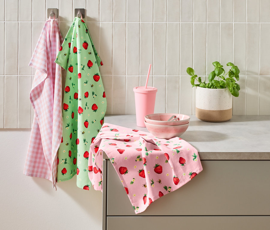 Na kuchyňské lince stojí tři utěrky, růžový hrnek s brčkem, dvě růžové misky a květináč bazalky.