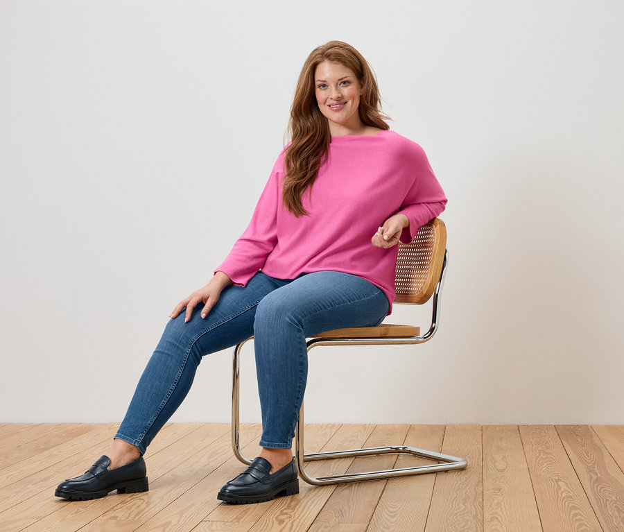Žena sedí na židli a má na sobě růžový svetr, modré džíny a černé mokasíny.