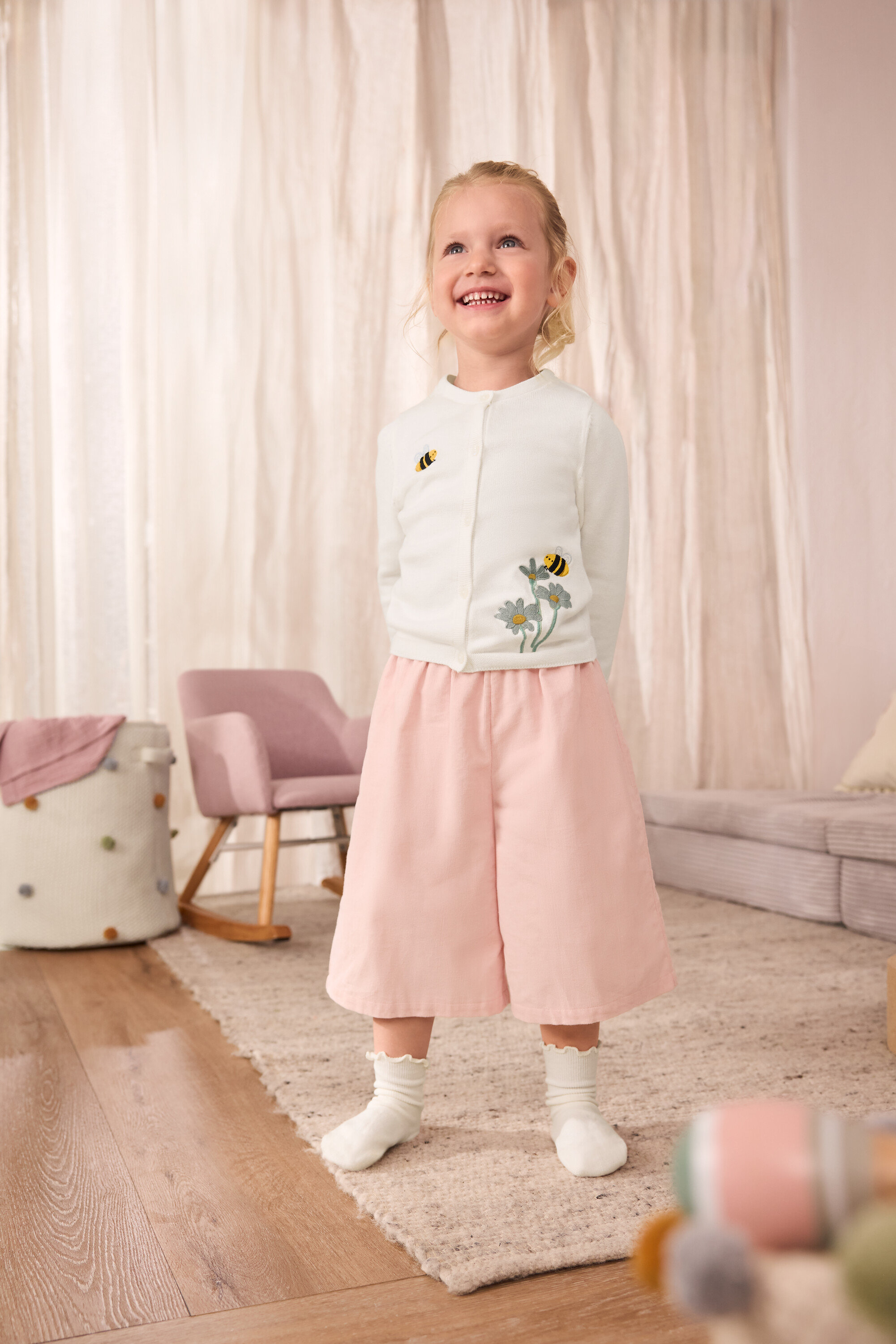 Usmívající se dívka má na sobě dívčí pletený svetr s vyšitými včelami, dětské manšestrové kalhoty culotte a dívčí ponožky s volánkovými lemy, 5 párů.