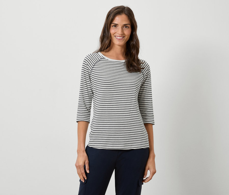 Žena má na sobě pruhované tričko s dlouhým rukávem a tmavě modré kalhoty na bílém pozadí.