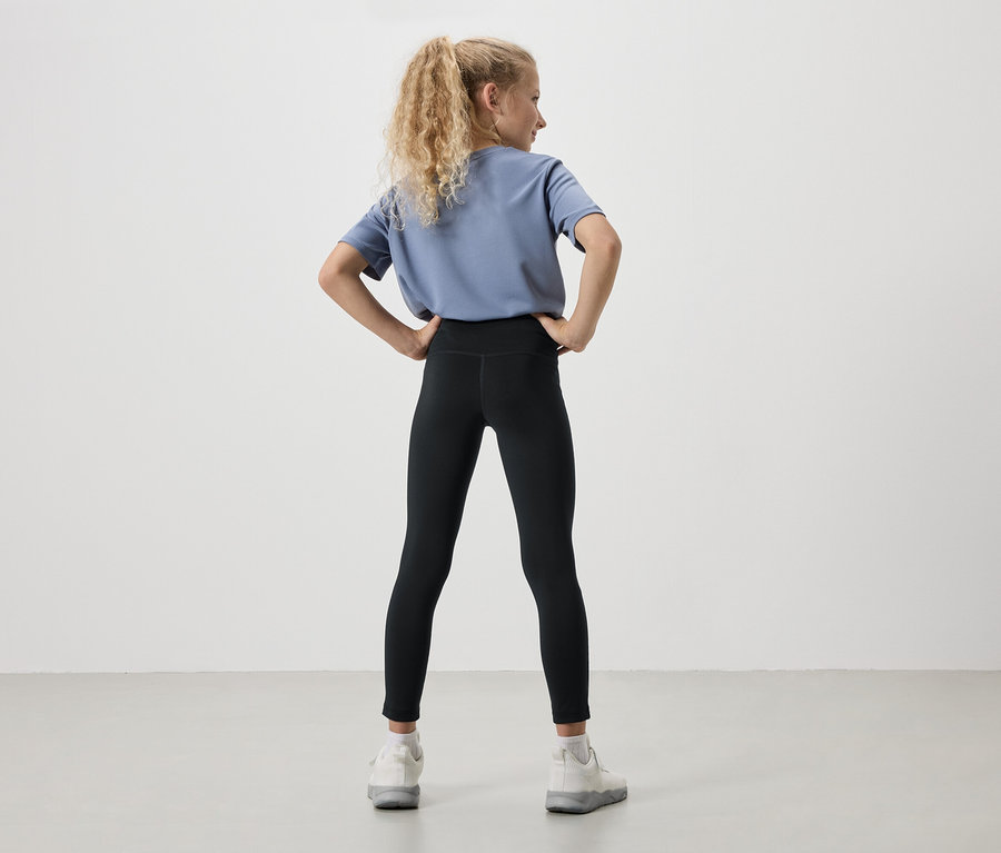 Pohled zezadu na mladou dívku s blond kudrnatými vlasy, oblečenou v modrém tričku, černých legínách a bílých teniskách, s rukama v bok.