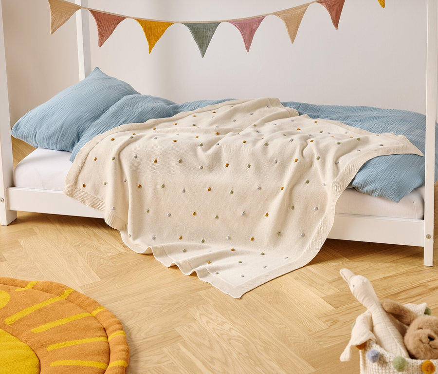 Dětská postel s modrými polštáři a krémovou dekou s barevnými puntíky stojí na dřevěné podlaze. Nad postelí visí barevný praporkový banner.