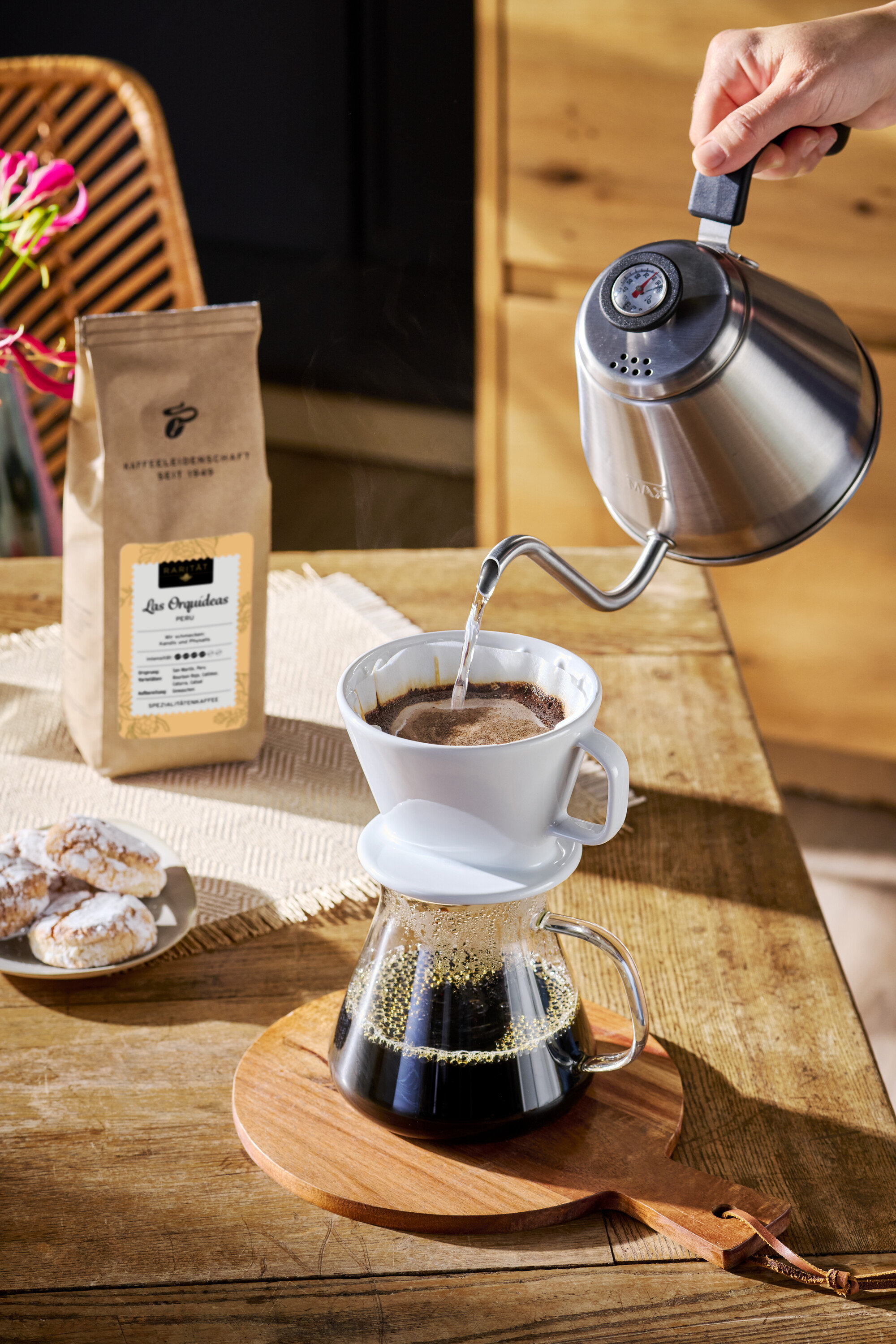 Ruka nalévá vodu ze stříbrné konvice do bílého kávového filtru. Vedle stojí balíček kávy a talíř sušenek.