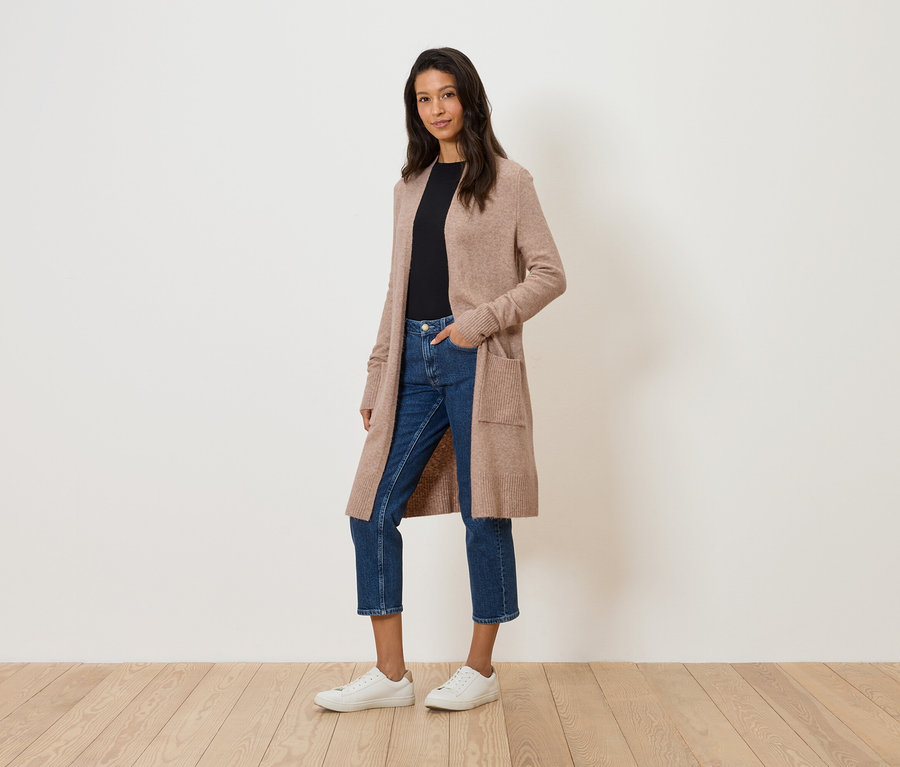 Žena stojí na bílém pozadí v dlouhém béžovém kardiganu, džínách a bílých teniskách.