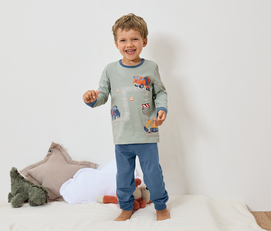 Usmívající se chlapec stojí bosý na bílém povrchu a má na sobě šedé pyžamo s modrými manžetami a motivem bagru a míchačky betonu.