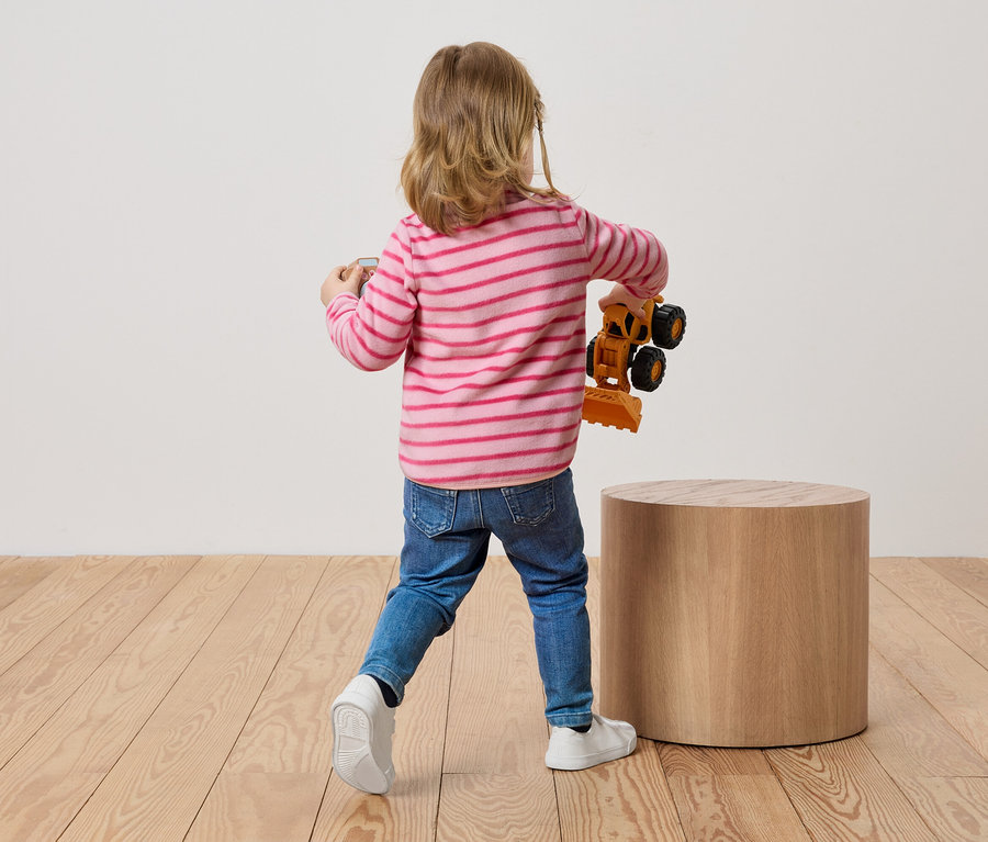 Dítě s blond vlasy stojí zády k divákovi a drží v ruce hračku bagru.