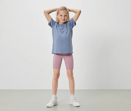 Dívka s blond vlasy stojí se zvednutýma rukama, má na sobě modré tričko, růžové cyklistické šortky a bílé tenisky.