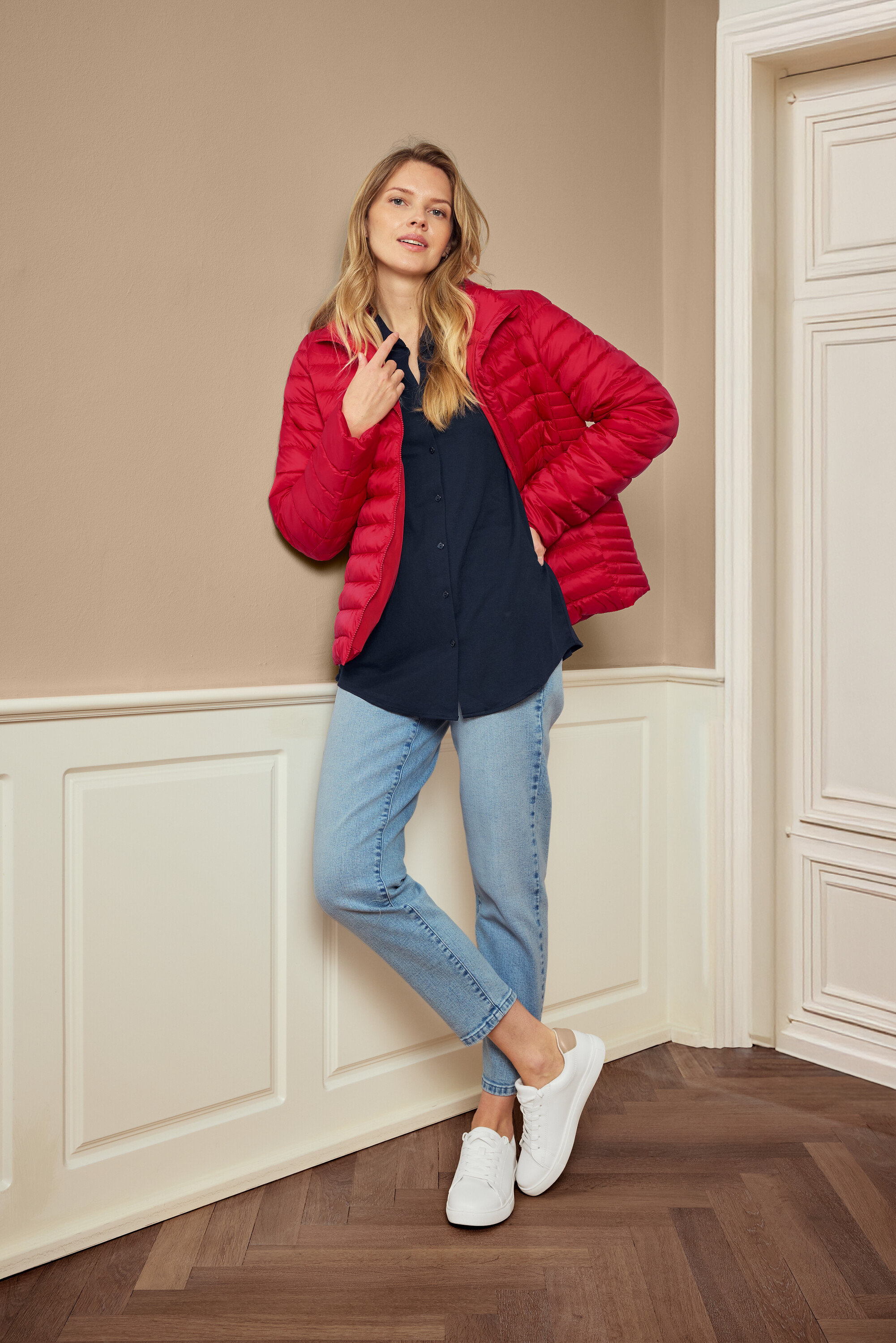 Žena pózuje v červené bundě, modré košili, džínách a bílých teniskách, má na sobě náušnice 925 Silber Creolen Hexagon.