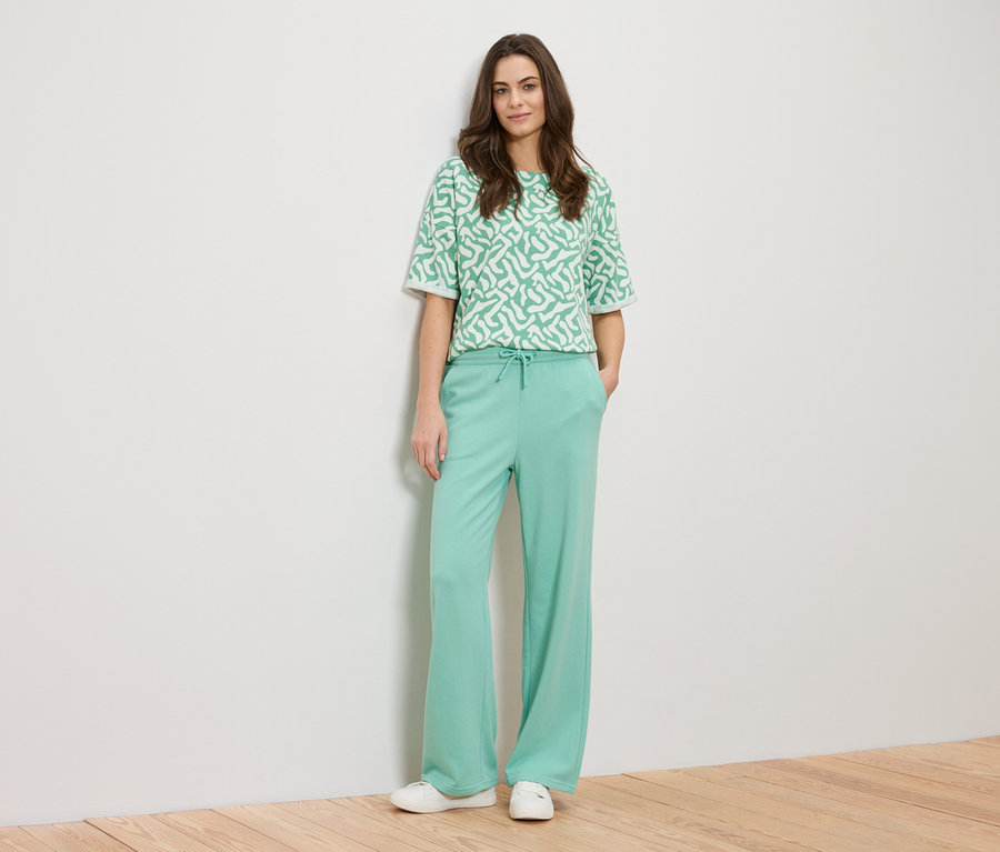 Žena se opírá o zeď a má na sobě zelený vzorovaný top a zelené kalhoty.