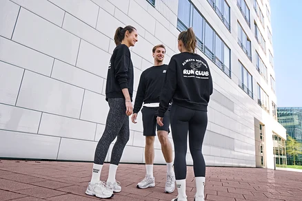 Tři lidé, dvě ženy a jeden muž, stojí usměvaví a povídají si. Všichni mají na sobě černé mikiny a bílé unisex tenisové ponožky Coffee Crew. Muž má černé sportovní šortky. Jedna žena má šedo-černé sportovní legíny »ActivePower«, druhá černé sportovní legíny »ActivePower«.
