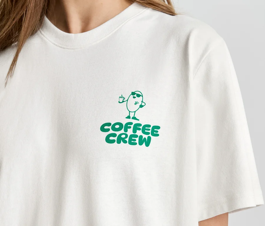Detail osoby v světlém tričku se zeleným potiskem "Coffee Crew" a kreslenou postavičkou kávového zrna.