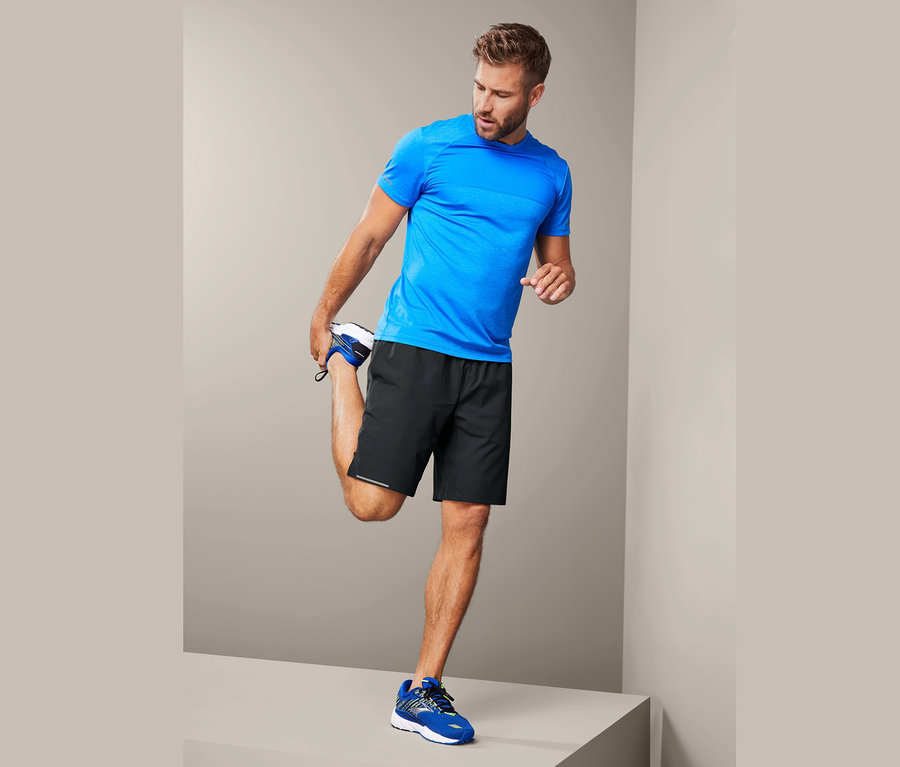 Muž protahuje nohu na šedém pozadí. Má na sobě modré tričko a černé sportovní šortky.