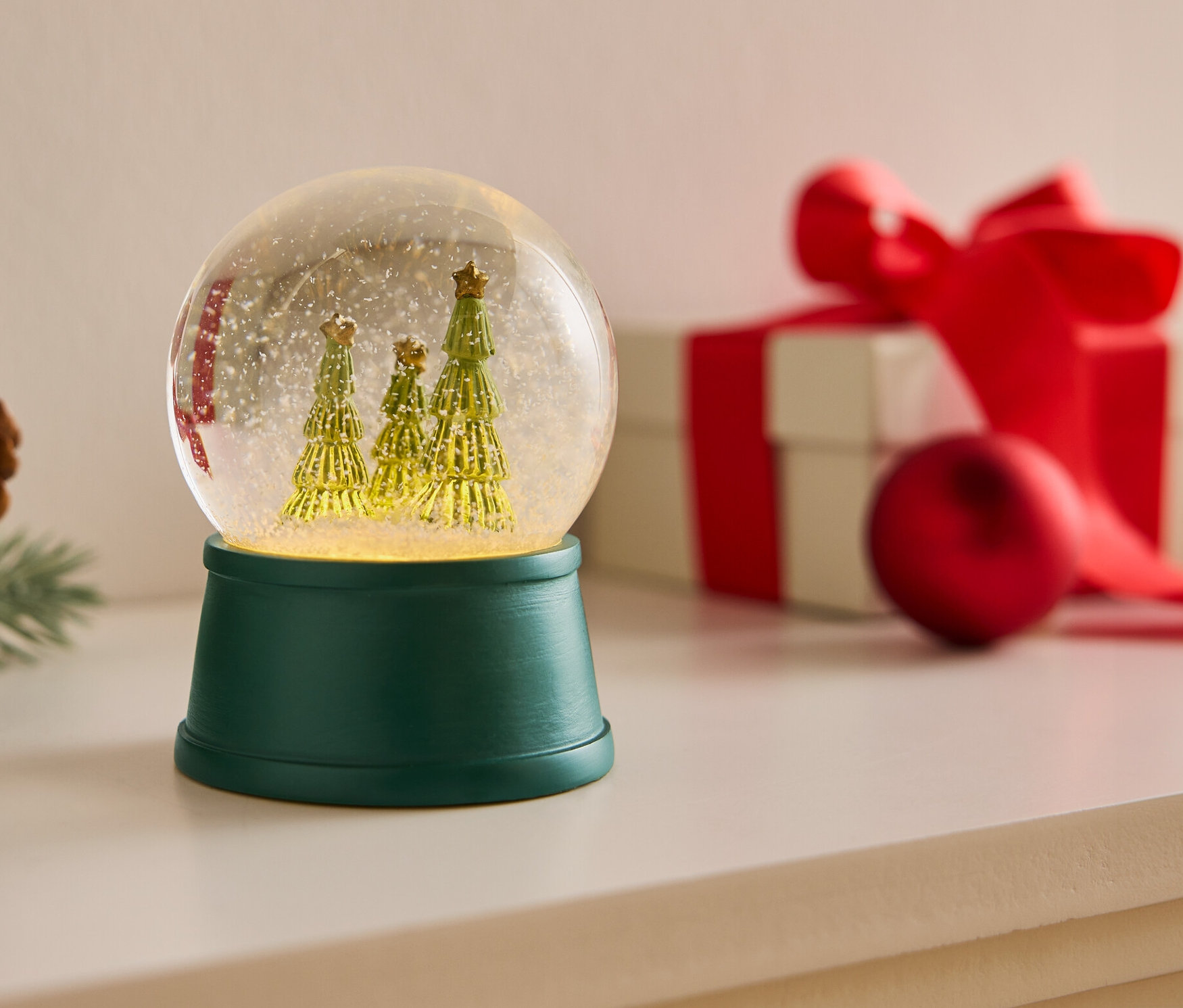 Sněhová koule se třemi stromy na zeleném podstavci vedle šišky a dárku s červenou stuhou.