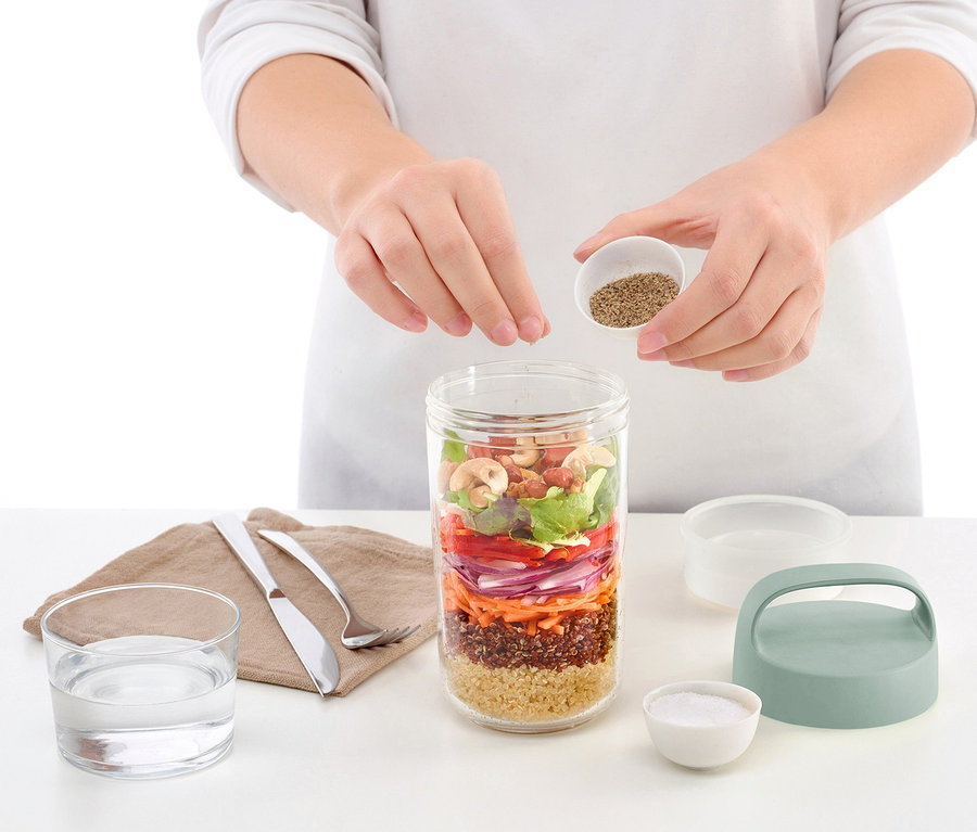 Žena sype koření do cestovní dózy na oběd, která je plná vrstev salátu.