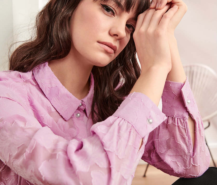 Žena s hnědými vlasy má ruce u obličeje a na sobě má světle fialovou košilovou halenku se strukturovaným vzorem.
